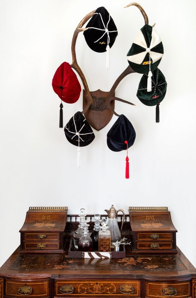 Antique wooden desk beneath mounted antlers displaying traditional embroidered velvet sports caps.