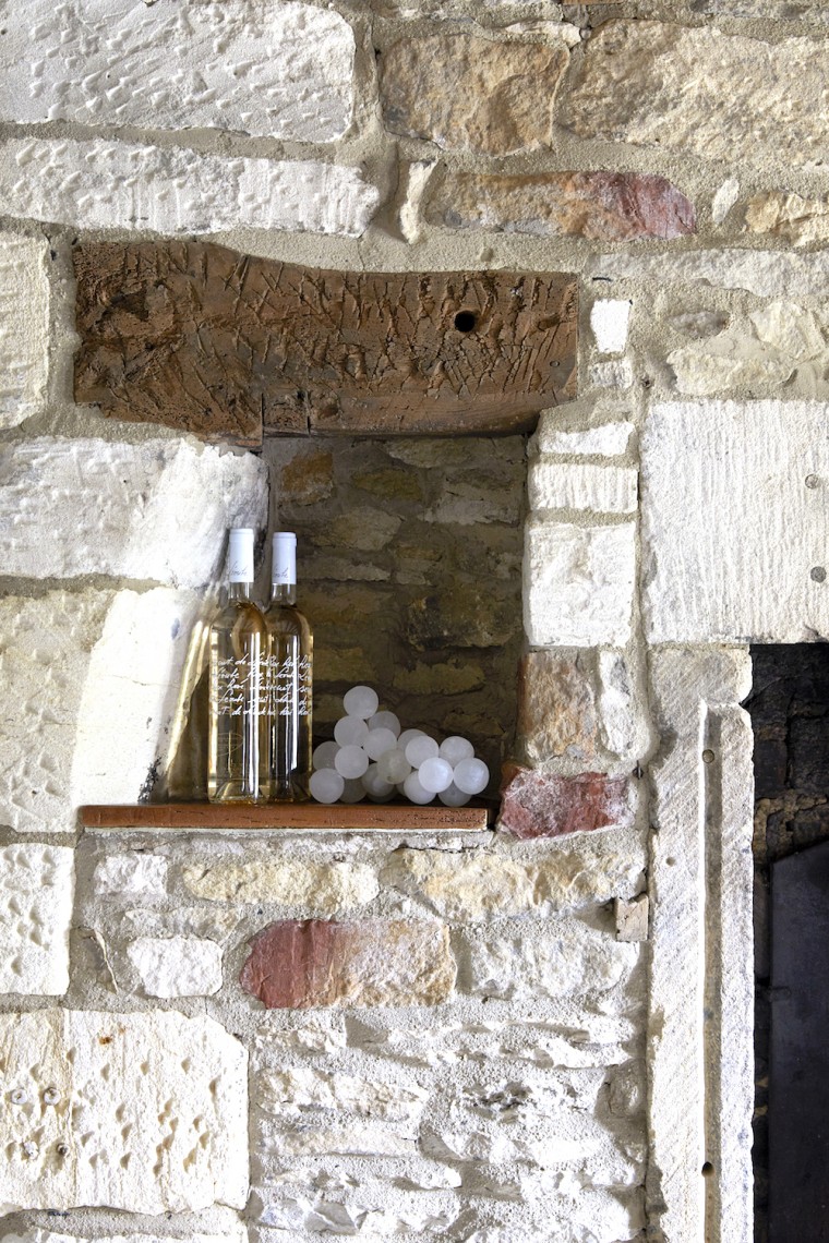 Recessed stone wall niche with aged timber lintel, styled with glass bottles and decorative spheres in a Cotswold country cottage.
