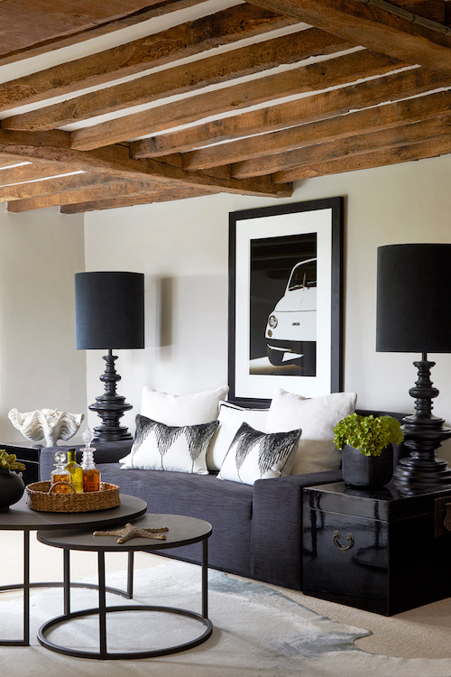 Cotswold country cottage sitting room with exposed timber beams, tailored dark sofa, monochrome cushions, and classic black table lamps creating a relaxed yet refined interior.