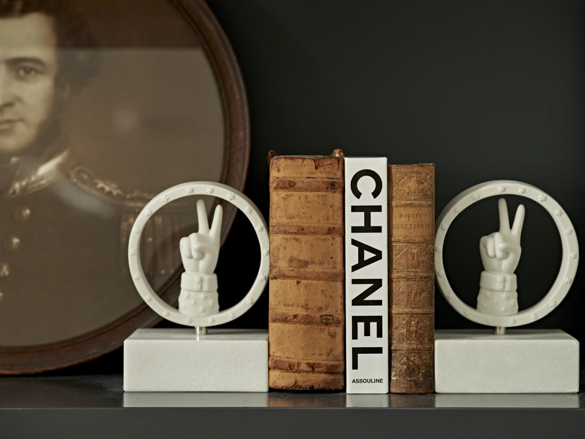 An Eclectic styled bookshelf vignette featuring antique leather-bound books, a white “Chanel” Assouline volume, sculptural bookends with hand motifs, and a vintage portrait backdrop.