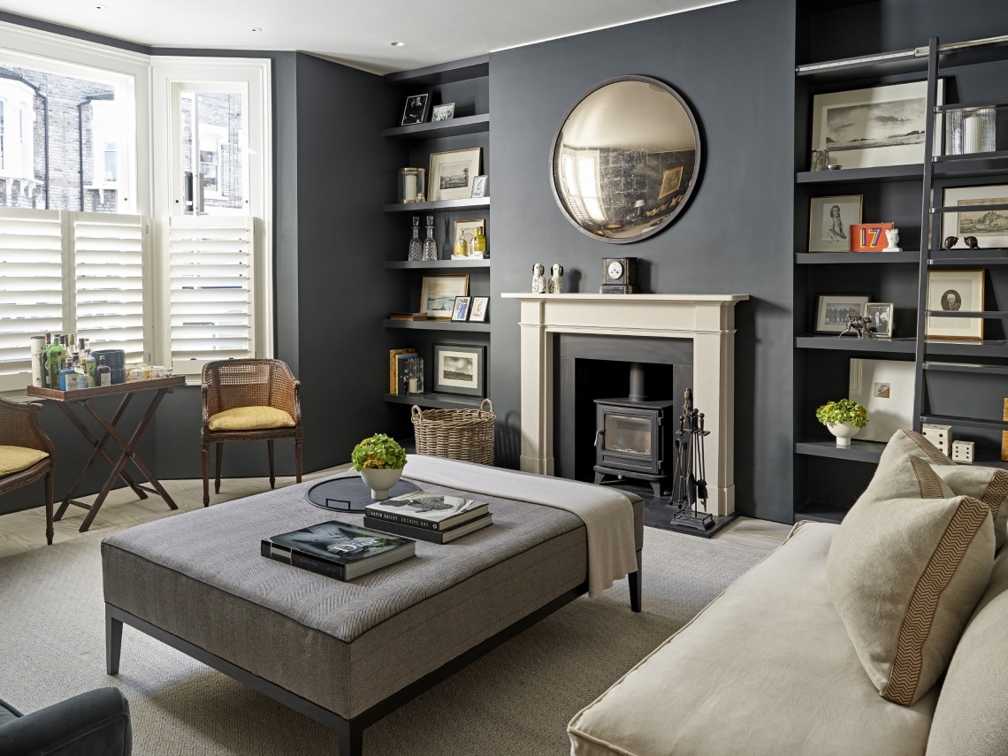 Refined London townhouse sitting room with deep grey walls, bespoke built-in shelving, a stone fireplace with wood-burning stove, round antique mirror and a central upholstered ottoman, styled with layered books and soft neutral furnishings.