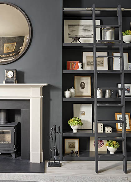 Dark-painted living room with bespoke built-in shelving and library ladder beside a stone fireplace, styled with framed artwork, books and decorative objects.