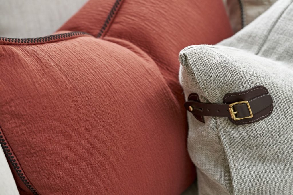 Close-up of layered sofa cushions in soft grey and warm rust tones, highlighting textured fabrics and a leather strap detail for a refined, country-inspired interior.