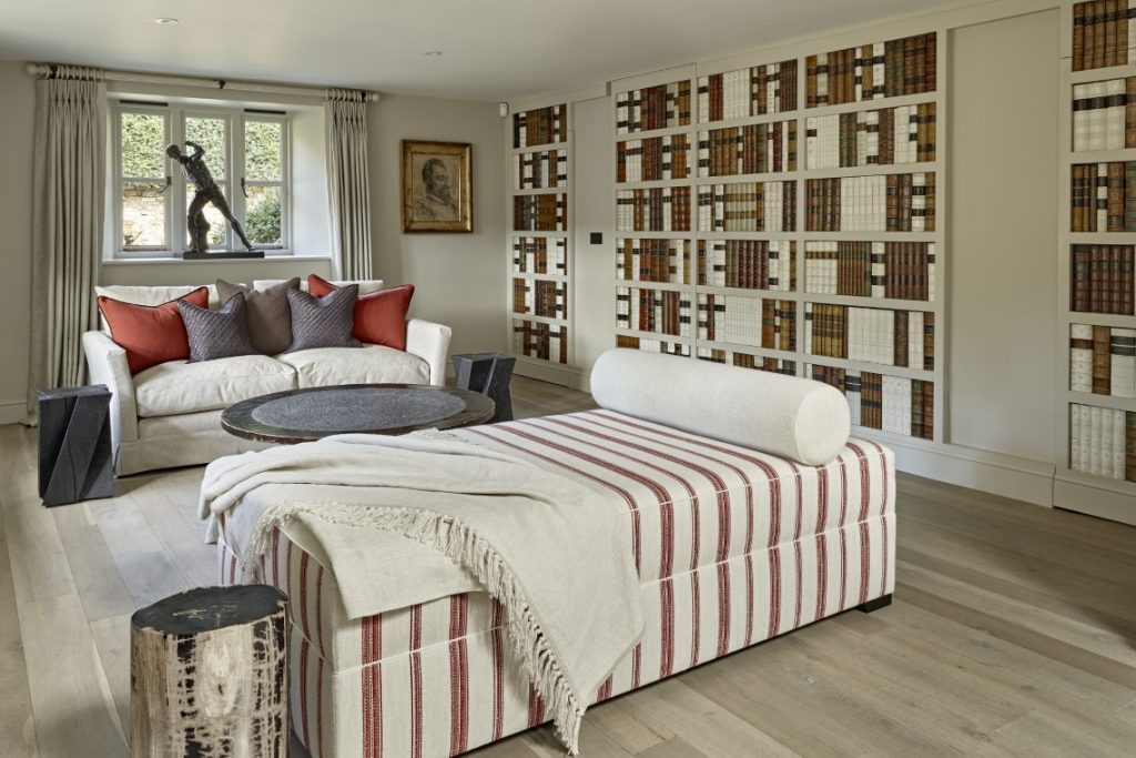 Living room with a bespoke false library wall of leather-bound book spines concealing a secret door, paired with a striped upholstered daybed, soft neutral seating and warm accent cushions for a refined country retreat feel.