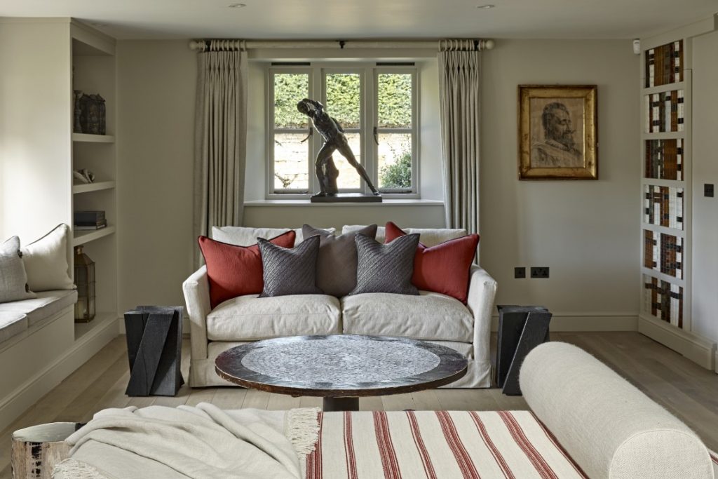 Living room within a Cotswold country retreat featuring a bespoke false library wall with concealed door, neutral upholstered sofa with rust and grey cushions, sculptural coffee table, and a window-seat sculpture framed by soft curtains.