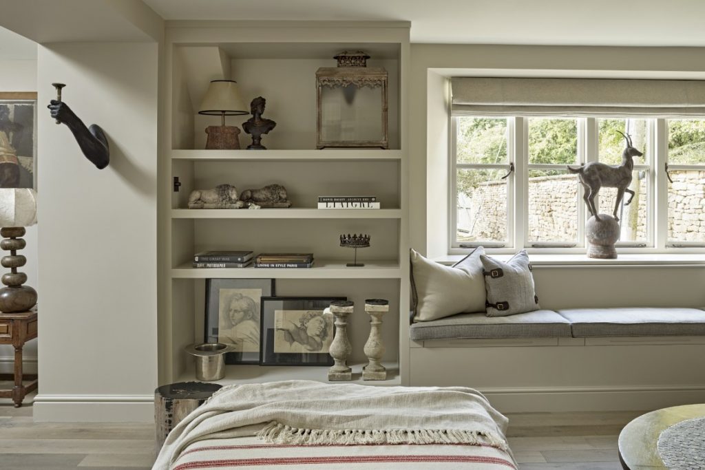 Built-in shelving and window seat in a Cotswold country retreat, styled with curated antiques, artwork and soft neutral cushions overlooking a stone garden wall.
