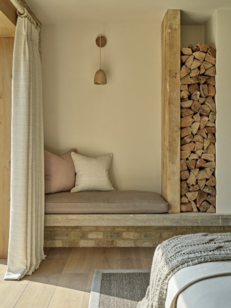 Calm seating nook within a converted Cotswold barn featuring natural materials, stacked firewood and soft neutral textiles.