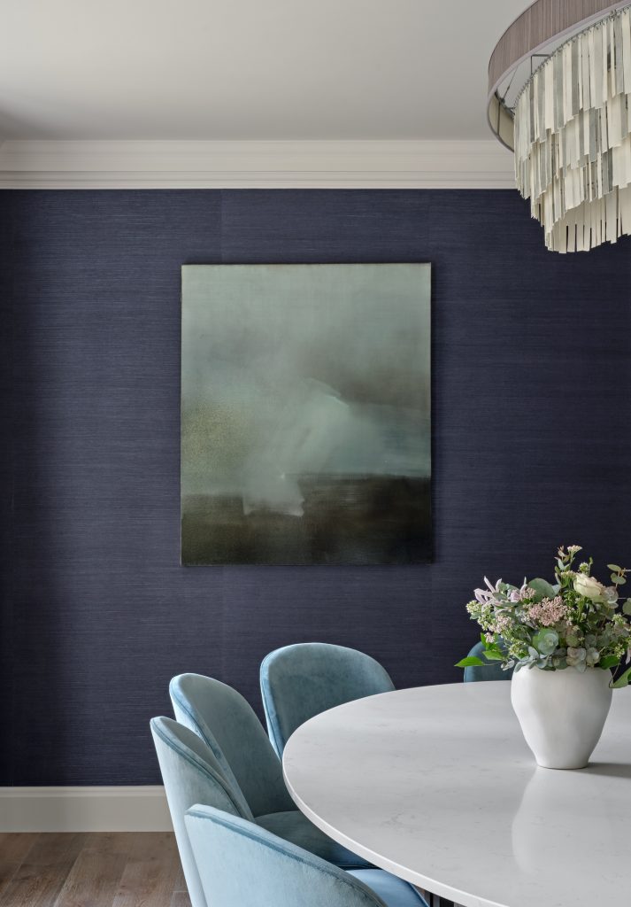 Dining room with deep blue textured walls, a sculptural chandelier and a bespoke oval dining table, softened with pale blue velvet chairs and contemporary artwork.