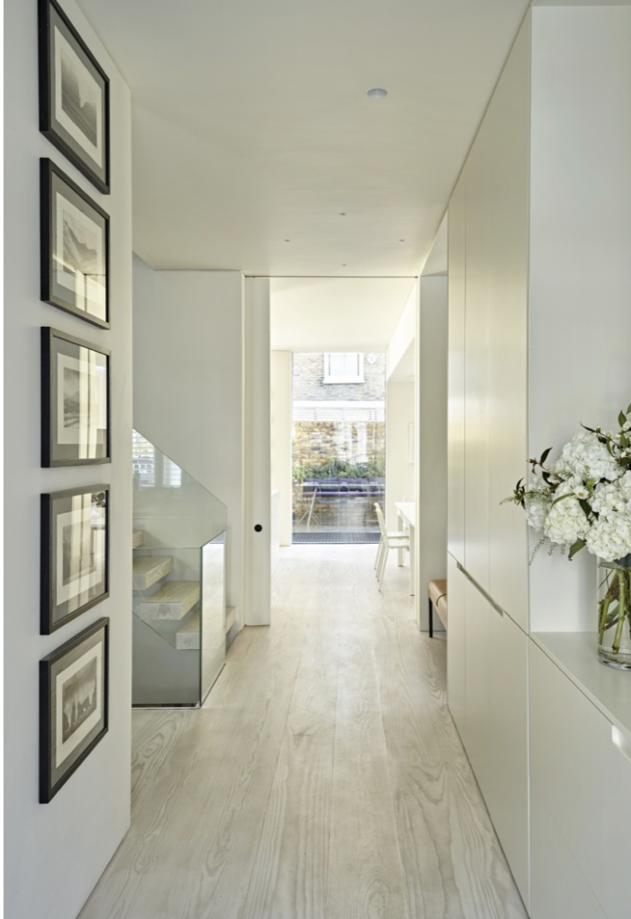 Light-filled entrance hall in a London family townhouse with pale oak flooring, bespoke built-in cabinetry, glass balustrade staircase and a monochrome gallery wall leading to the rear garden.