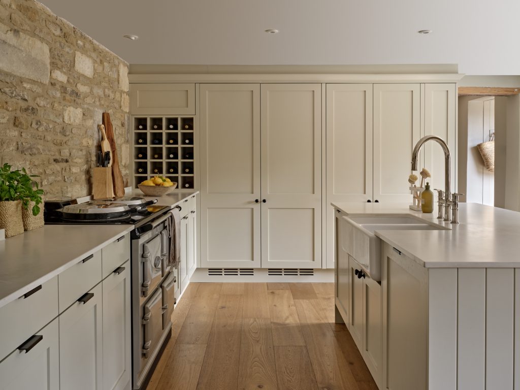 Cotswold country cottage kitchen with handcrafted shaker cabinetry, exposed stone wall, farmhouse range cooker, and a classic island with Belfast sink, finished with warm oak flooring.