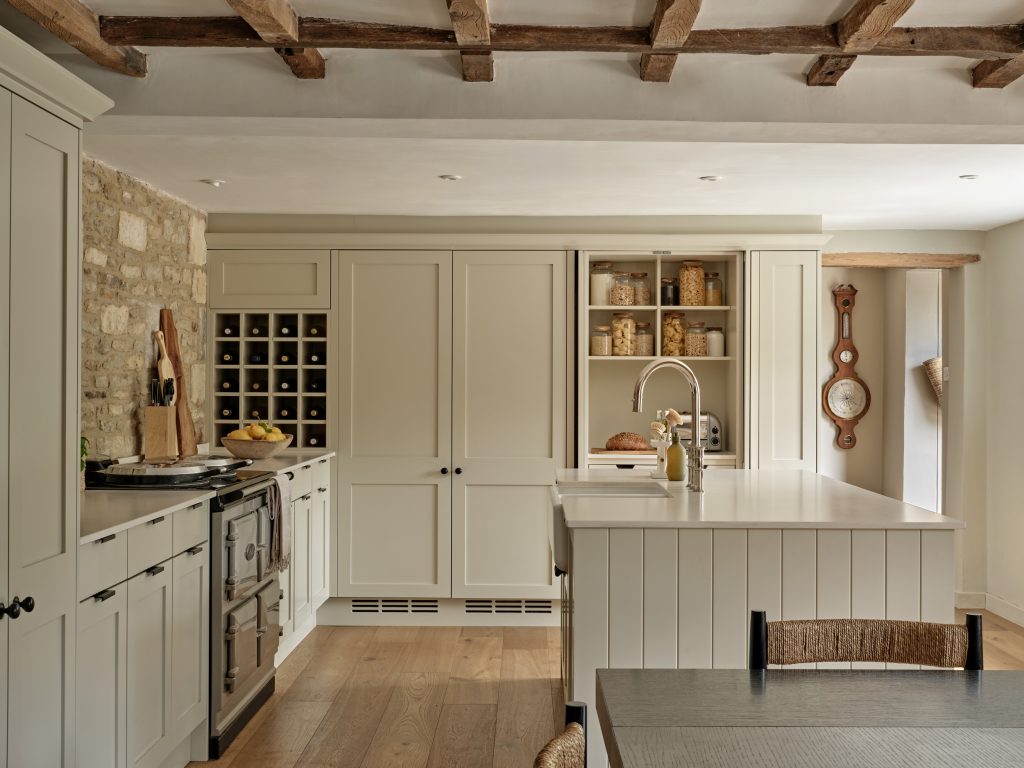 Bespoke Cotswold country cottage kitchen featuring painted shaker cabinetry, exposed oak beams, limestone wall detailing, a farmhouse range cooker and a generous island with Belfast sink, blending traditional craftsmanship with understated elegance.