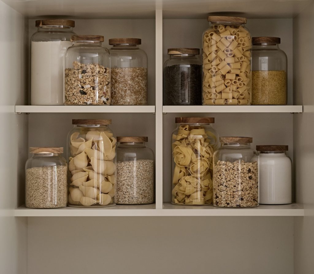 Neatly organised pantry shelving within a Cotswold country cottage kitchen, featuring glass storage jars with wooden lids filled with pasta, grains, flour and cereals, creating a calm, practical and timeless farmhouse aesthetic.