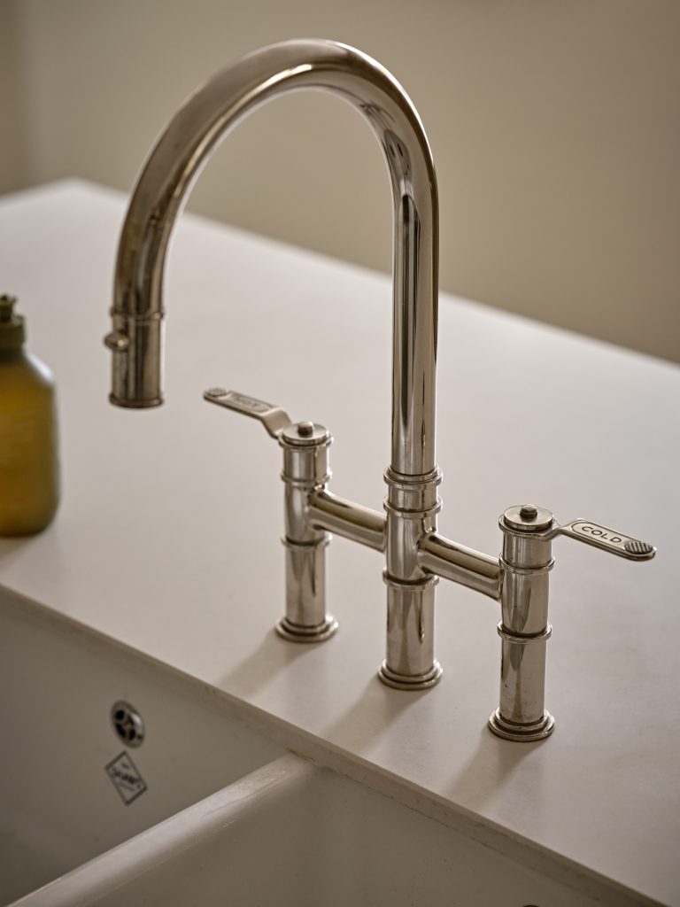 Close-up of a polished nickel bridge mixer tap over a Belfast sink, highlighting classic lever handles and refined detailing within a Cotswold country cottage kitchen.